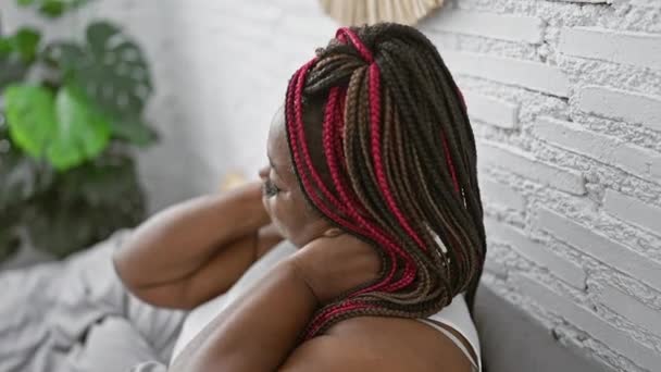 美丽的非洲裔美国女人 头戴辫子 患有颈痛 不幸地坐在阳光充足的卧室里舒适的床上 生动地描绘了她忧心忡忡的室内生活方式 — 图库视频影像