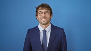 Takım elbiseli neşeli genç adam, gözlerini bir kolla kapatıyor, eğlenceli bir şekilde gülümsüyor, gözleri bağlı bir saklambaç gibi bir ifadeyle, soğuk, izole edilmiş mavi bir arka plan üzerinde..