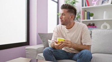 Kendine güvenen genç İspanyol adam sabah espresso 'sunun keyfini çıkarıyor, oturma odasındaki kanepesinde rahatça oturuyor, yüzünde ışıl ışıl bir gülümseme çiçek açıyor.