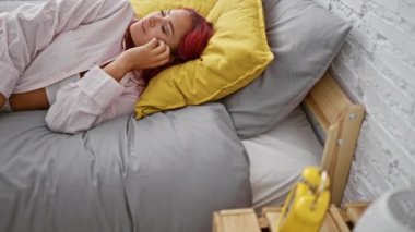 Sıcak bir sabah, güzel, gevşemiş kızıl saçlı yetişkin yatakta rahat bir şekilde uzanıyor, dikkatle çalar saate bakıyor, evin içindeki yatak odasının yumuşak ışığında zamana konsantre oluyor.