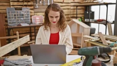 Gülümseyen genç kadın marangoz, çevrimiçi marangozluk işine dalmış, iç mekandaki atölyesinde dizüstü bilgisayarla bir masada oturuyor.