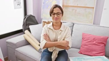 Canlı genç İspanyol kadın sanatçı, stüdyodaki koltukta çapraz silahlarla oturuyor, pozitiflik yayıyor, başarıyı ve güveni somutlaştırıyor.