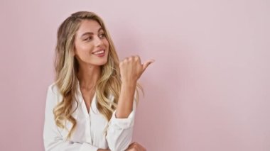 Genç sarışın kadın, tişört giyip gülümsüyor ve gülümsüyor. Gülümsüyor ve başparmağıyla yan tarafa bakıyor. izole edilmiş pembe arkaplan
