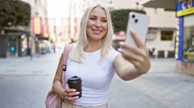 Güneşli bir şehir caddesinde, akıllı telefonuyla yaptığı bir video konuşmasında kendinden emin bir şekilde gülümseyen genç, sarışın bir kadın..