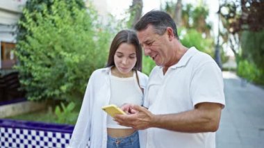 Mutlu İspanyol baba ve kızı parkta eğleniyor, ayakta dururken ve akıllı telefon kullanırken birlikte gülümsüyorlar.