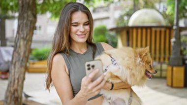 Genç, kendine güvenen İspanyol kadın neşeyle gülümsüyor, güzel köpeğine sarılıyor, parkta telefonuyla neşeli bir şekilde mesajlaşıyor..