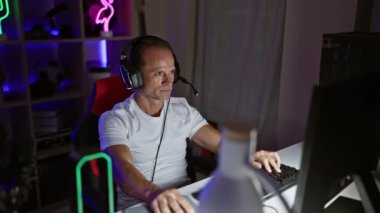 Neşeli orta yaşlı adam, muzaffer flamalı, oyun odasında kulaklıklarını sevinçle çıkarıyor, destansı bir video oyununun galibiyetini işaret ediyor.