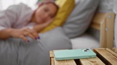 Uyanık ve ciddi, güzel kızıl saçlı bir kadın, rahat bir şekilde dinleniyor, yatakta uzanıyor, rahat yatak odasında akıllı telefon saatine bakıyor, sabah saatine konsantre oluyor..