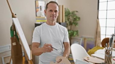 Yaratıcılıktan zevk alan orta yaşlı İspanyol sanatçı, kolej sanat stüdyosunun sıcak atmosferinin ortasında palet ve boya fırçasıyla rahat bir poz veriyor.