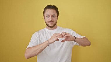 Young hispanic man smiling confident doing heart gesture with hands over isolated yellow background