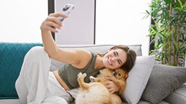 Neşeli, genç İspanyol kadın evinde, oturma odasındaki kanepesinde rahatça gülümsüyor. Evcil köpeğiyle mutlu bir selfie çekmek için akıllı telefon teknolojisi kullanıyor..