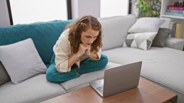Oturma odasındaki kanepede oturan odaklanmış genç bir kadın dikkatle evdeki dizüstü bilgisayarını kullanıyor. Kafkasyalı güzel bir yetişkinin ciddi bir portresi. Cihazının üzerinde. Kapalı alandaki işine dalmış..