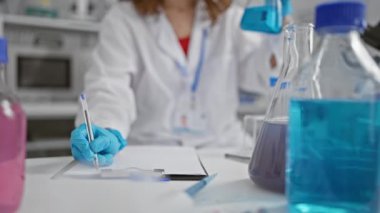 Test tüpündeki sıvıyı analiz eden odaklanmış genç kadın bilim adamı verimli modern laboratuvarda notlar alıyor.