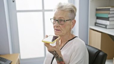 Ofiste çalışan beyaz saçlı çekici yaşlı kadın zarif bir iş gülüşüyle otururken akıllı telefonundan sesli mesaj yolluyor.