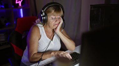 Çok sıkılmış, orta yaşlı sarışın flamalı kadın oyun odasında video oyunu oynuyor, gece yarısı deresinden yorgun düşmüş.