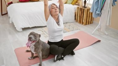 Sportif, gri saçlı, orta yaşlı, evcil köpeği olan bir kadın. Yatak odasının sakin atmosferinde yoga eğitimi alıyor. Sağlıklı egzersiz rejimi için yatağın yanında oturuyor.