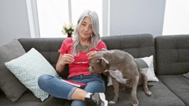 Kendinden emin orta yaşlı bir kadın, gri saçlı köpeğiyle, kanepede oturup edebiyat okumaktan zevk alıyor, evinde neşe ve rahatlık ifade ediyor..
