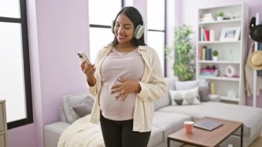 Kaygısız genç İspanyol kadın, beklenen anneliğin sevinciyle parıldıyor, neşeli bir şekilde dans ediyor ve karnına dokunuyor. Oturma odasında akıllı telefonuyla müzik dinliyor.