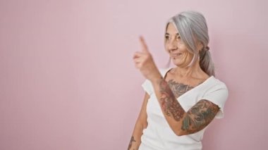 Kendine güveni tam, olgun bir kadın pembe arka planda sevinçle yan tarafı işaret ediyor.