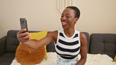 Neşeli Afro-Amerikalı kadın evinde rahat koltuğunda canlı bir video görüşmesi yapıyor. Yüzü ışıl ışıl gülüyor..