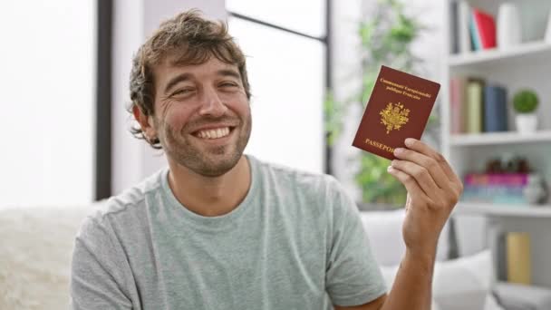 ジョイフルな若い男は 彼の家のソファーに気軽に座って フランスのパスポートを保持しながら自信を持って微笑んで 次のヨーロッパ旅行の準備 — ストック動画