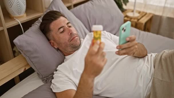 苦恼的年轻人穿着睡衣躺在床上 焦急地看着智能手机 同时在卧室里拿着一瓶药丸 — 图库视频影像