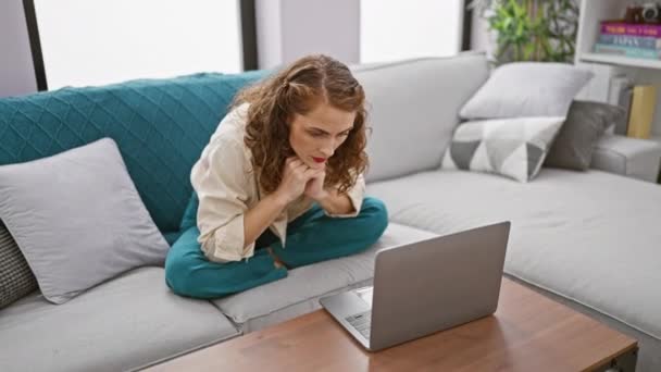 专注的年轻女性坐在客厅沙发上 专注地在家里使用笔记本电脑技术 一个漂亮而集中的高加索成年人的严肃的肖像在她的装置上 全神贯注于她在室内的工作 — 图库视频影像
