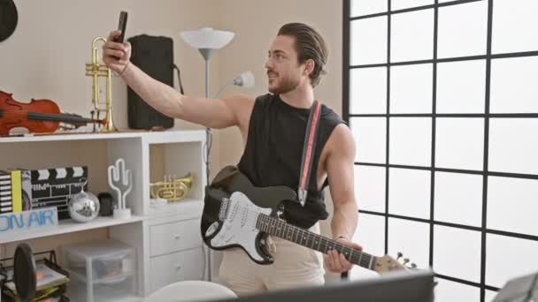 音楽スタジオでスマートフォンでセルフィーを作る電気ギターを演奏する若いヒスパニックな男ミュージシャン — ストック動画