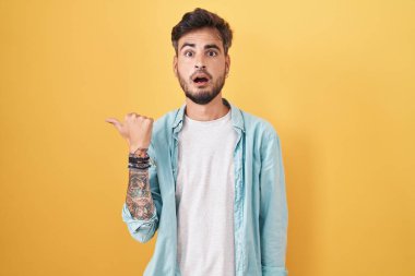 Sarı arka planda duran İspanyol dövmeli genç adam elleri yana dönük, ağzı açık şaşkın bir ifadeyle şaşırdı.. 