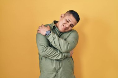 Sarı arka planda duran İspanyol genç adam kendine sarılıyor mutlu ve pozitif, kendinden emin gülümsüyor. Sevgi ve özgüven. 