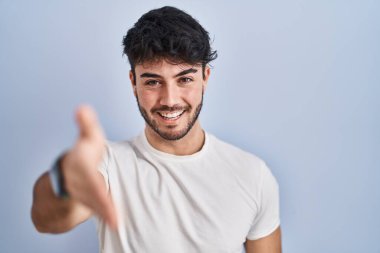 Hispanic man with beard standing over white background smiling friendly offering handshake as greeting and welcoming. successful business. 