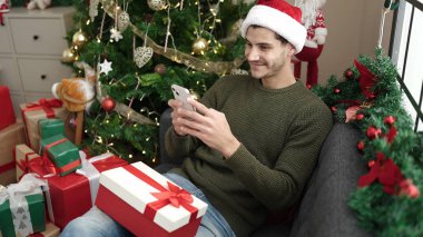 Young hispanic man using smartphone sitting on sofa by christmas tree at home