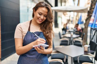 Young beautiful hispanic woman waitress smiling confident writing on notebook at coffee shop terrace