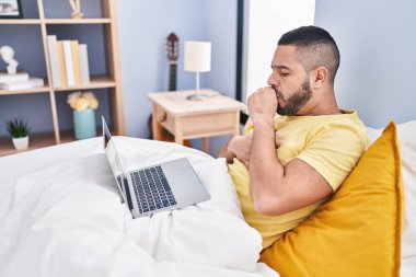Young latin man coughing having telemedicine sitting on bed at bedroom