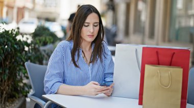 Genç ve güzel İspanyol kadın akıllı telefon kullanıyor kahve dükkanının terasında alışveriş torbalarıyla oturuyor.