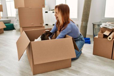Beyaz bir kadın yeni evinde elinde karton kutunun köpeğiyle yerde oturuyor.