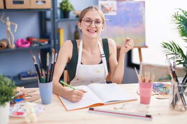 Genç, beyaz, öğrenci üniforması giyen bir kadın sanat paletini kullanarak masada oturuyor ve gülümsüyor. 