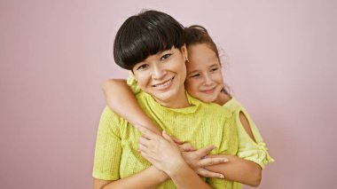 Kendine güvenen anne ve kız, mutlu bir kucaklaşmayı paylaşırken izole edilmiş pembe bir arka planda birlikte duruyorlar. Keyif, neşe ve pozitif titreşimlerle dolu sıradan bir yaşam tarzını ifade ediyorlar..