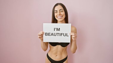 Küstah sabah enerjisi! Genç, kendine güvenen, İspanyol bir kadın, seksi bir şekilde iç çamaşırıyla, elinde 'Ben güzelim' afişi, izole edilmiş pembe bir arka planda saç stiliyle hava atıyor..
