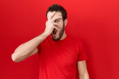 Gündelik kırmızı tişört giyen genç İspanyol adam şok içinde yüzünü ve gözlerini kapayarak utangaç ifadesiyle parmaklarının arasından bakıyor.. 
