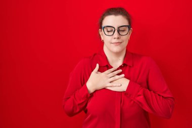 Kızıl arka planda duran, elleri göğsünde, gözleri kapalı ve yüzünde minnet dolu jestlerle gülümseyen Latin kökenli genç bir kadın. sağlık kavramı. 
