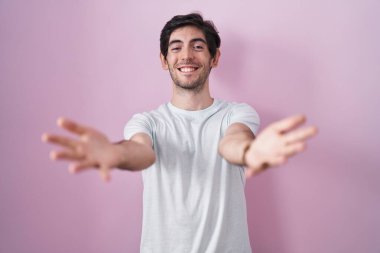 Young hispanic man standing over pink background looking at the camera smiling with open arms for hug. cheerful expression embracing happiness. 