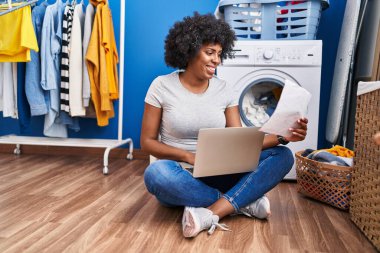 Afrika kökenli Amerikalı bir kadın çamaşır odasında çamaşır makinesini beklerken dizüstü bilgisayar okuyor.