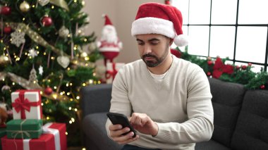Young hispanic man using smartphone sitting on sofa by christmas tree at home