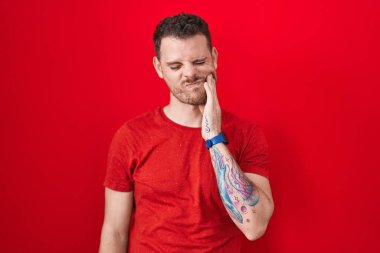 Kırmızı arka planda dikilen İspanyol genç adam diş ağrısı ya da diş ağrısı yüzünden acı dolu bir ifadeyle ağıza dokunuyor. dişçi 