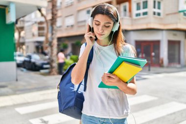 Young beautiful hispanic woman student holding books using smartphone at street