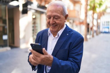 Orta yaşlı, kır saçlı, kendine güvenen, caddede akıllı telefon kullanan bir adam.