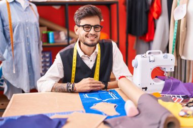 Young hispanic man tailor smiling confident sitting on table at atelier