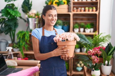 Young beautiful hispanic woman florist smiling confident holding plant at florist