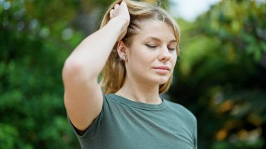 Parkta saçlarını tarayan ciddi bir ifadeyle yan tarafa bakan genç sarışın kadın.